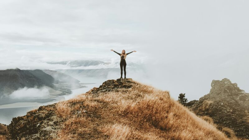 A woman stands with arms outstretched on a mountain ridge overlooking a lake and clouds.