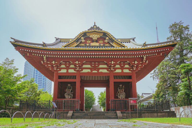 A red Japanese temple gate with golden ornaments and guardian statues, with skyscrapers and Tokyo Tower in the background.