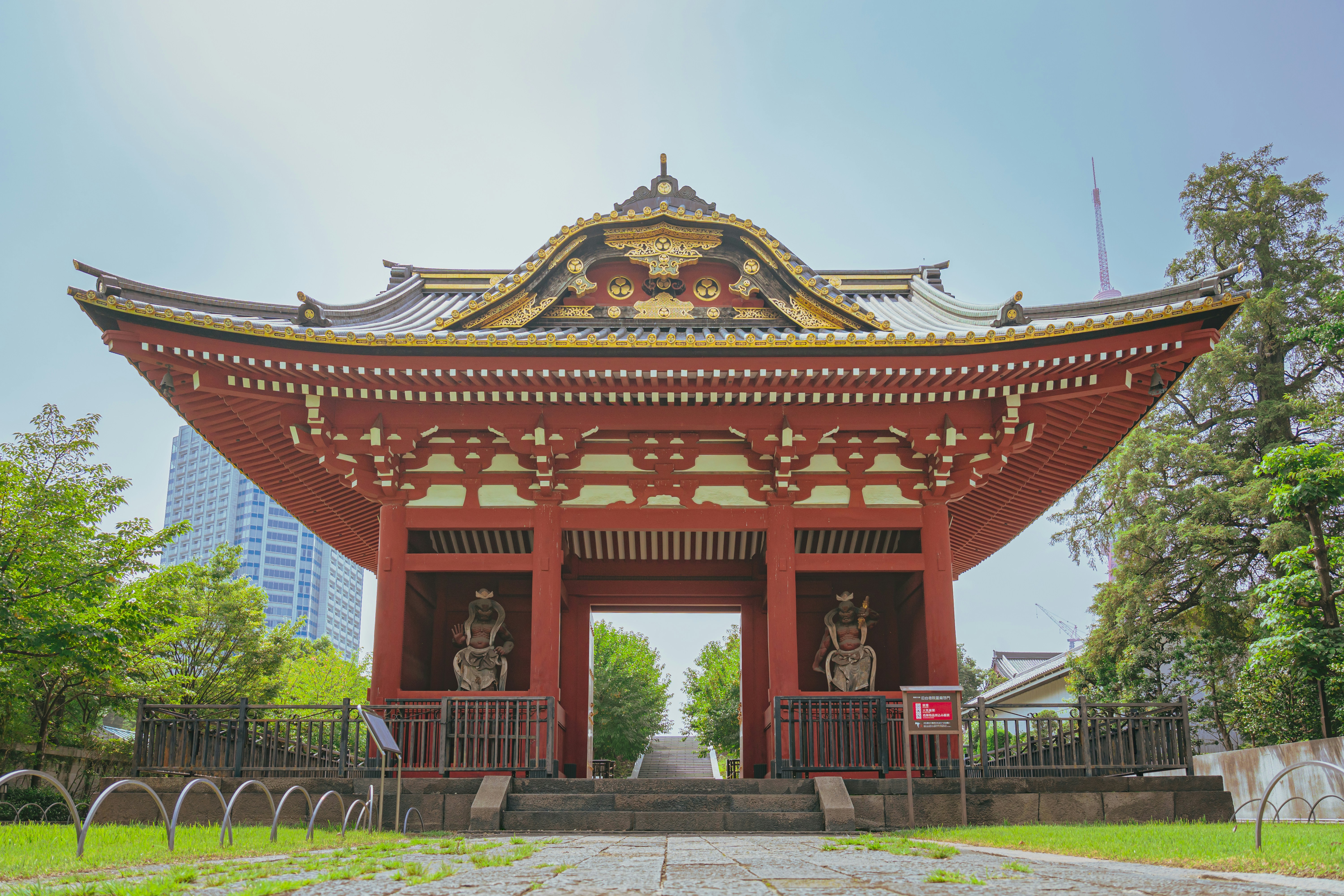 Červená japonská chrámová brána se zlatými ornamenty a sochami strážců, v pozadí mrakodrapy a Tokyo Tower.