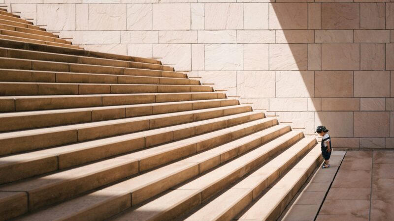 A small child in a cap stands at the foot of a tall stone staircase, looking down.